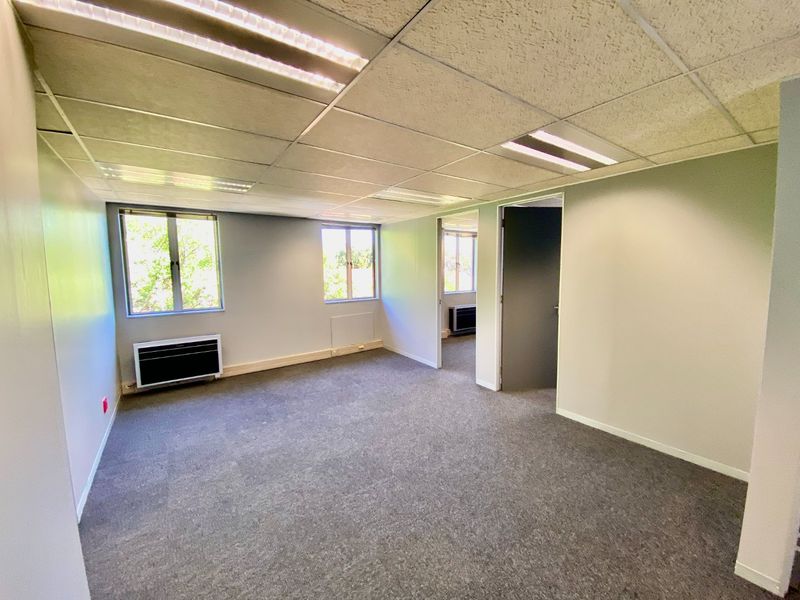 An empty commercial office space featuring carpeted flooring, a suspended ceiling with recessed lighting, and wall-mounted air conditioning units.