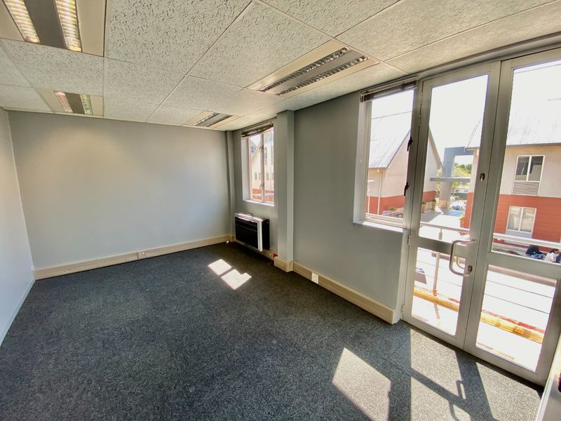 A bright, empty office space featuring carpeted flooring, a suspended ceiling with recessed lighting, and direct access to an exterior balcony through glass doors.