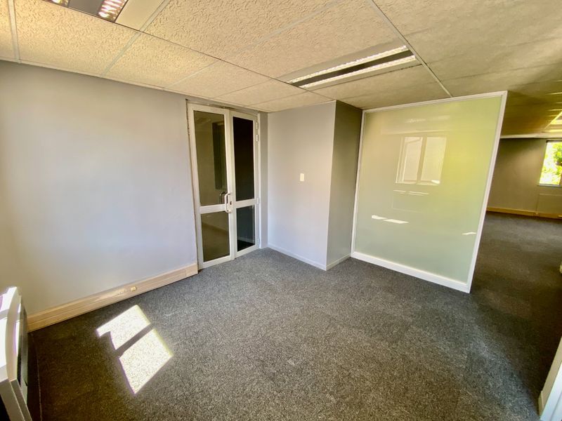 A vacant commercial office space featuring grey carpeting, a suspended ceiling with recessed lighting, and a modern frosted glass partition wall.