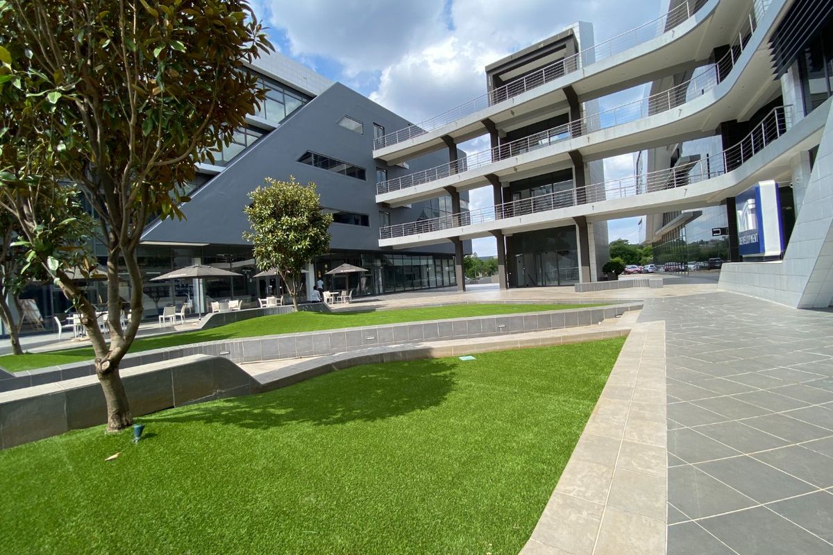 Modern commercial office complex featuring multi-level glass structures, connecting steel walkways, and a well-maintained landscaped courtyard with artificial turf.