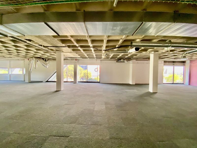 A large, open-plan commercial office space featuring an exposed waffle slab ceiling, industrial ductwork, carpet tiles, and expansive windows providing natural light.