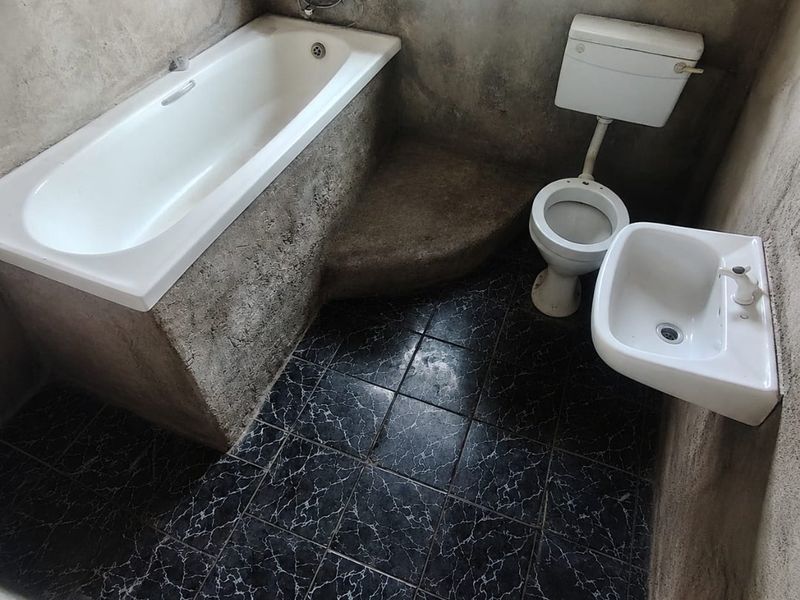 A bathroom featuring a built-in bathtub, toilet, and wall-mounted basin with dark tiled flooring and unfinished cement plaster walls.