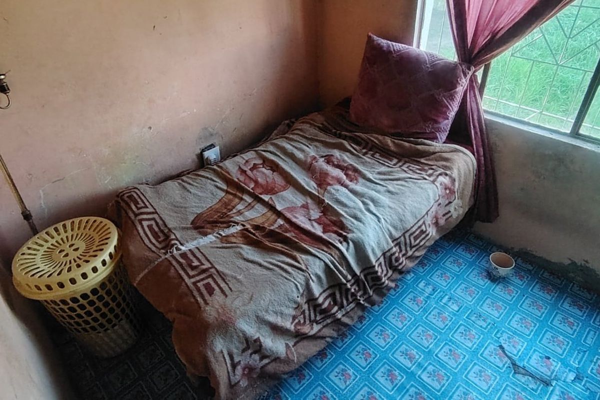 A small bedroom featuring tiled flooring and security bars on the window, currently showing significant wear to wall finishes and flooring.