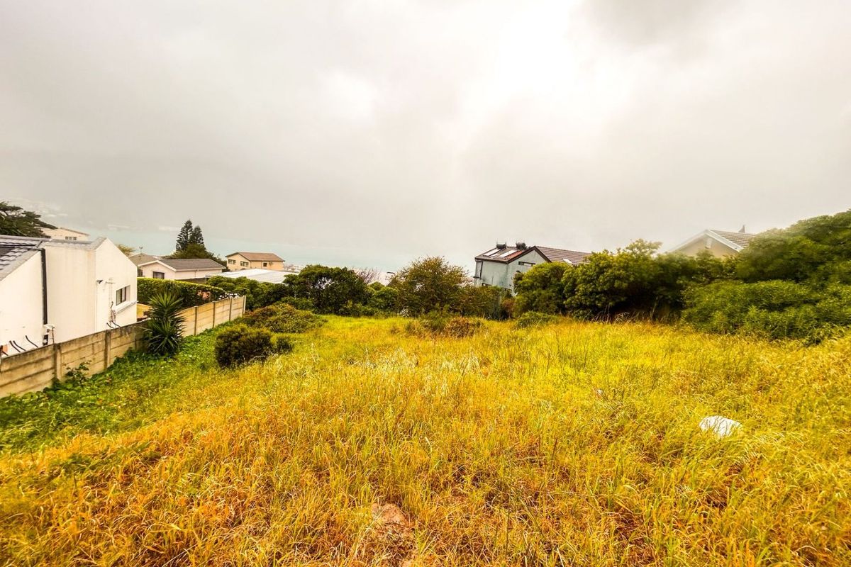 A vacant residential plot featuring overgrown grass and boundary walling, situated in a developed neighborhood with distant coastal views.