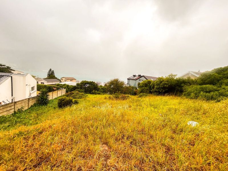 A vacant residential plot featuring overgrown grass and boundary walling, situated in a developed neighborhood with distant coastal views.