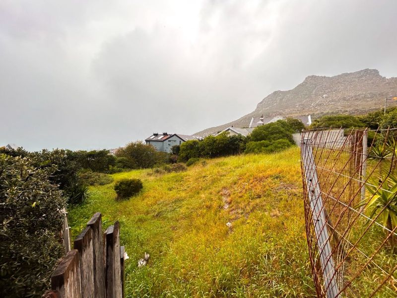 A view of a sloped, undeveloped residential plot featuring natural vegetation, enclosed by weathered wooden and rusted wire fencing with mountain views.