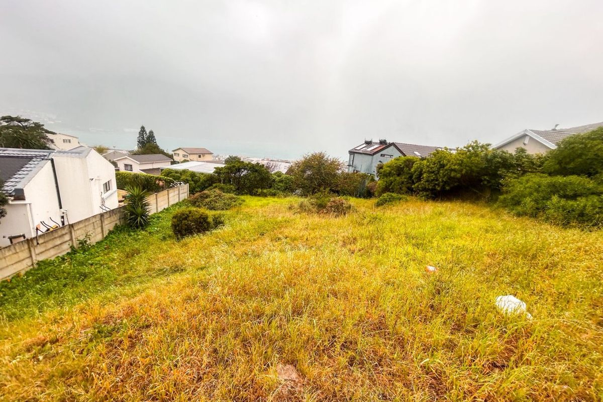 A sloped, undeveloped residential plot featuring overgrown grass and boundary walls, offering a view of the surrounding neighborhood and distant landscape.