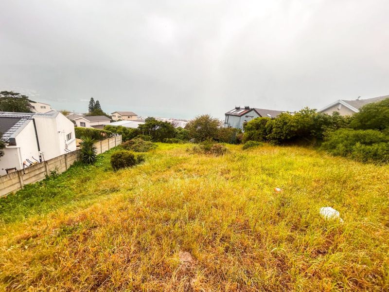 A sloped, undeveloped residential plot featuring overgrown grass and boundary walls, offering a view of the surrounding neighborhood and distant landscape.