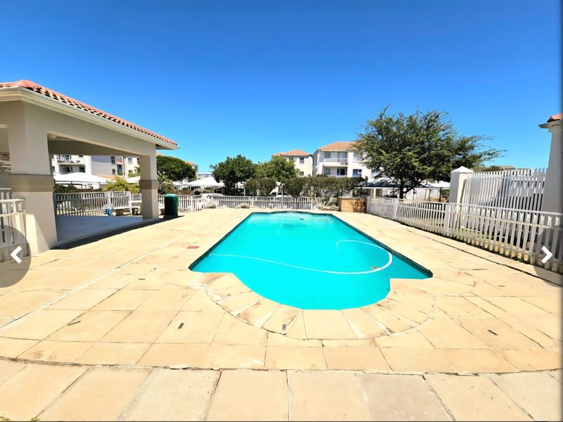 Communal swimming pool area featuring a paved deck, covered patio space, and perimeter fencing within an apartment complex.
