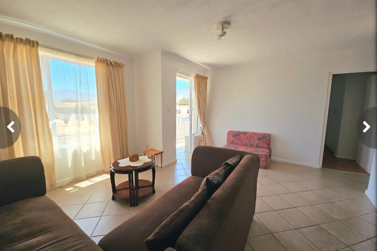 A bright living room featuring tiled flooring, large windows allowing natural light, and direct access to an outdoor area through a sliding glass door.