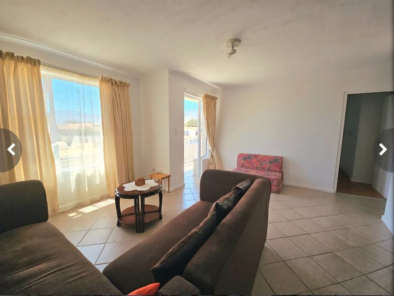 A bright living room featuring tiled flooring, large windows allowing natural light, and direct access to an outdoor area through a sliding glass door.