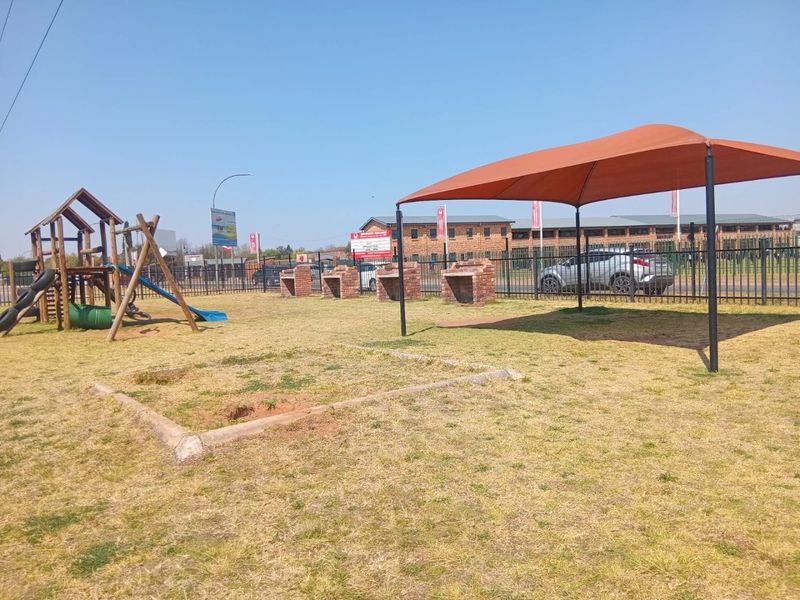 Communal garden area featuring brick braai stands, a shade sail structure, and playground equipment, enclosed by metal perimeter fencing.