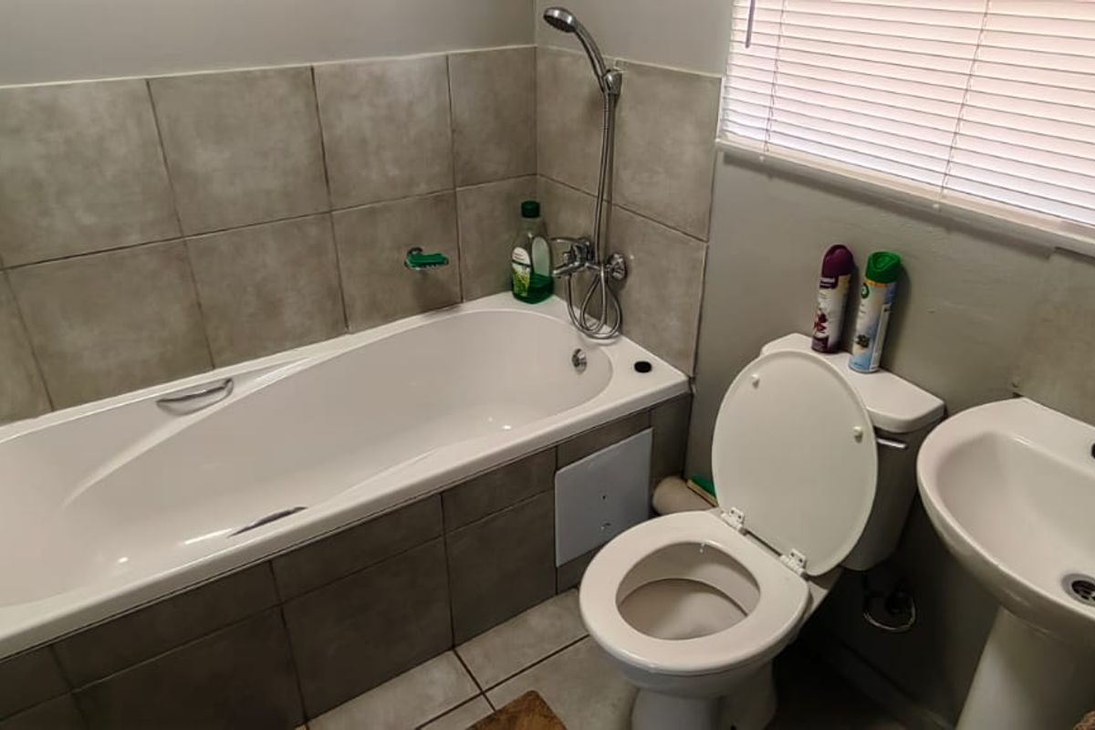 A functional bathroom featuring a bathtub with shower mixer, pedestal basin, and toilet, finished with neutral wall tiling and venetian blinds.