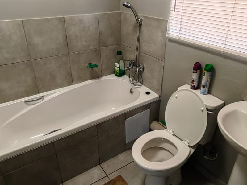 A functional bathroom featuring a bathtub with shower mixer, pedestal basin, and toilet, finished with neutral wall tiling and venetian blinds.
