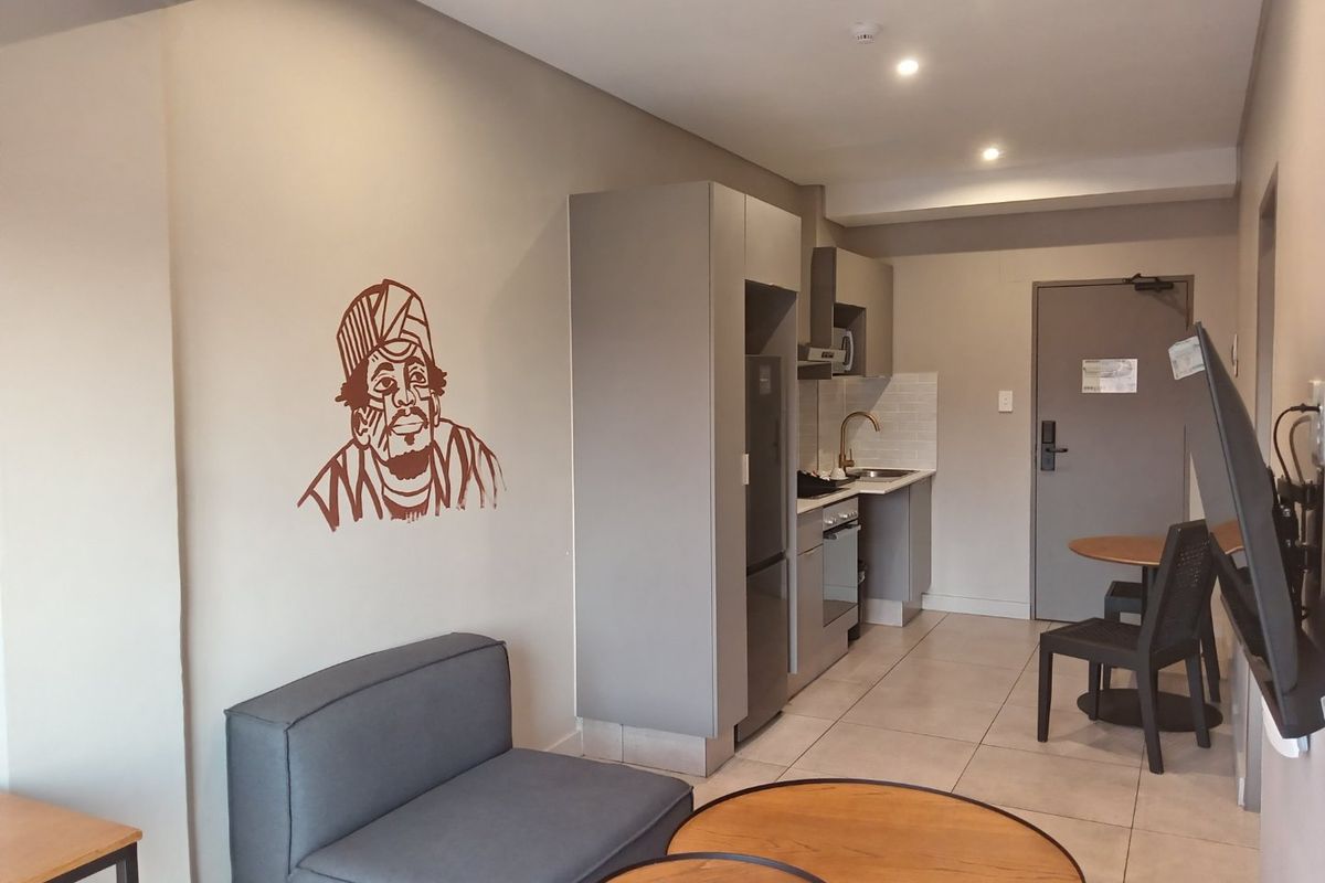 Open-plan apartment living area featuring a compact kitchenette with built-in cabinetry, integrated appliances, and neutral tiled flooring throughout.
