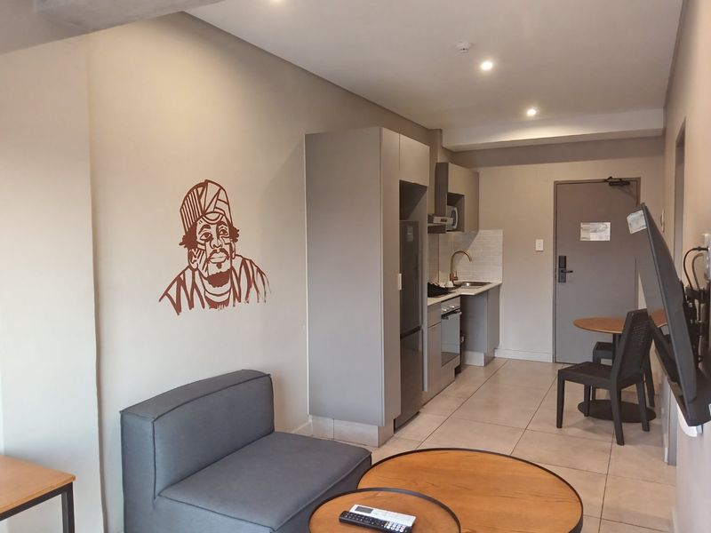 Open-plan apartment living area featuring a compact kitchenette with built-in cabinetry, integrated appliances, and neutral tiled flooring throughout.