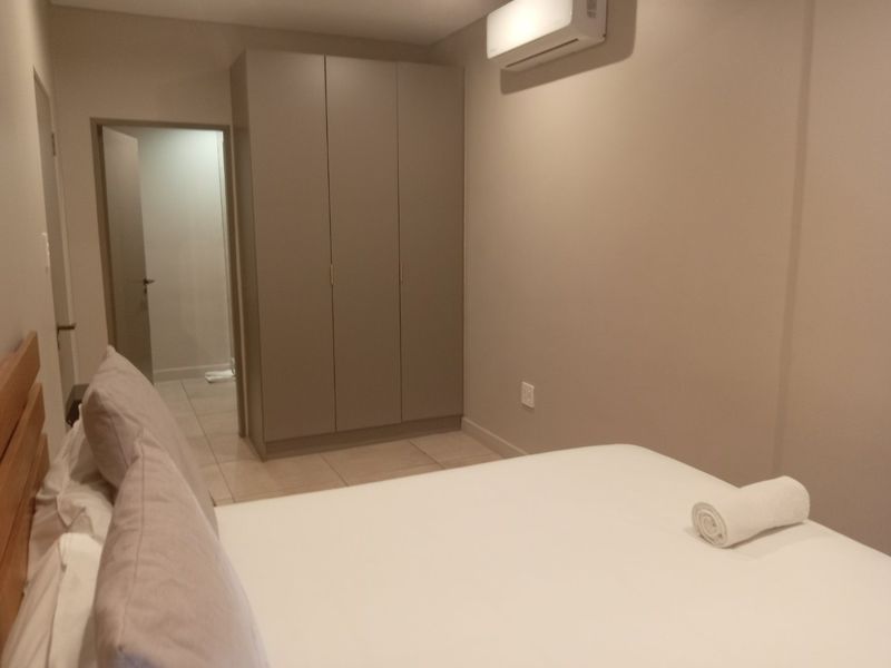 A clean, modern bedroom featuring neutral tones, tiled flooring, a built-in wardrobe, and a wall-mounted split-system air conditioning unit.