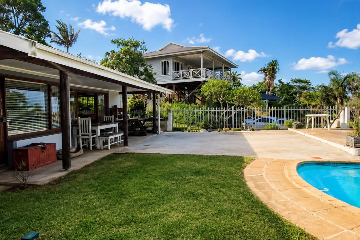Outdoor entertainment area featuring a swimming pool, concrete patio, and covered veranda, set within a landscaped garden with perimeter security fencing.