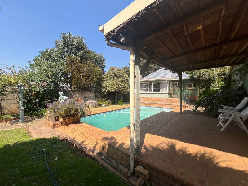 Outdoor pool area featuring a covered thatched patio, brick paving, and a swimming pool set within a landscaped garden.