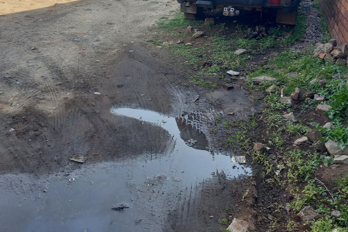 An unpaved residential front area showing poor drainage, muddy ground conditions, and a brick boundary wall.