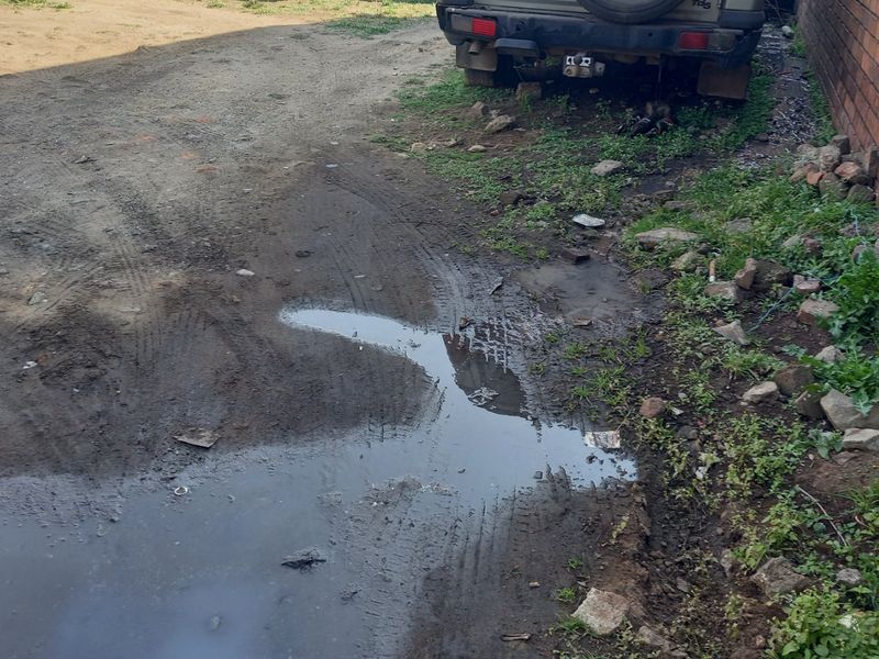 An unpaved residential front area showing poor drainage, muddy ground conditions, and a brick boundary wall.