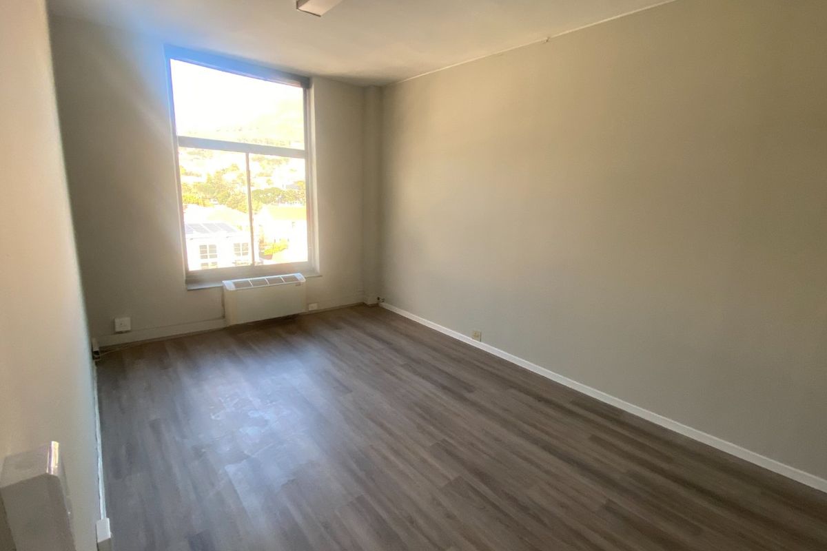 A bright, empty office space featuring laminate flooring, a large window with natural light, and a built-in HVAC unit.