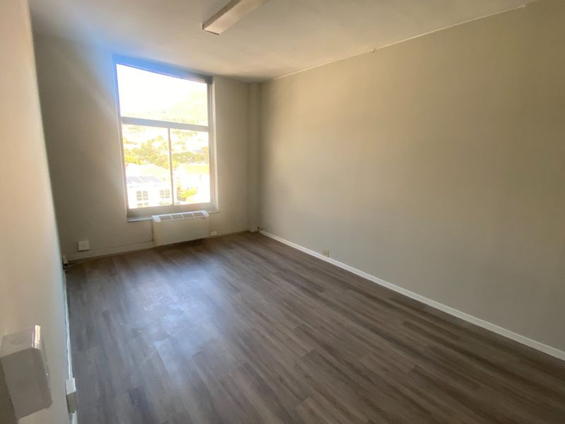A bright, empty office space featuring laminate flooring, a large window with natural light, and a built-in HVAC unit.