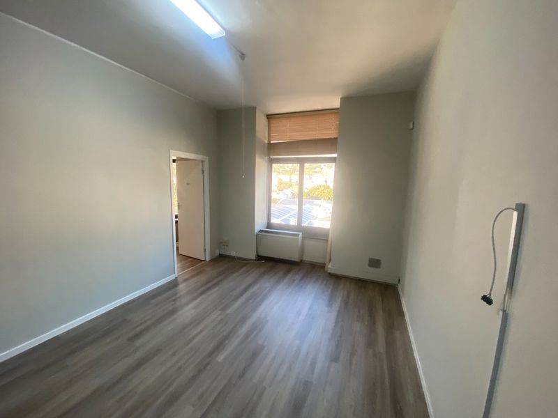 A bright, empty office space featuring modern laminate flooring, a built-in HVAC unit, and large windows with blinds.