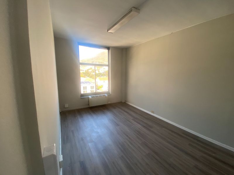 A bright, empty office space featuring laminate flooring, a large window with natural light, and a built-in HVAC unit.