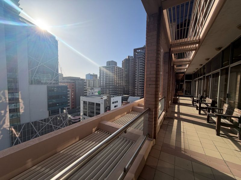 A spacious covered balcony featuring brick columns, tiled flooring, and a metal balustrade, offering an elevated urban view of the surrounding city skyline.