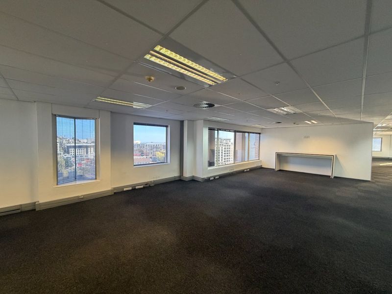 A spacious, vacant commercial office area featuring large windows with city views, suspended ceilings, recessed lighting, and dark carpet flooring.