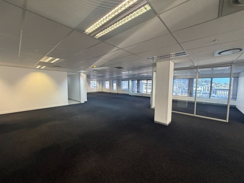 Spacious open-plan commercial office area featuring suspended ceilings, recessed lighting, dark carpeting, and glass partitioning with expansive city views.