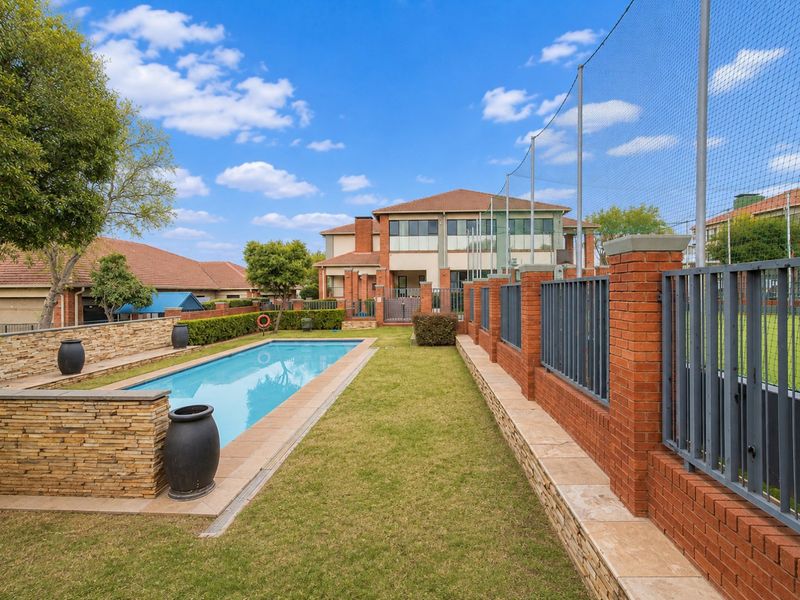 A well-maintained outdoor pool area featuring a rectangular swimming pool, manicured lawn, and secure brick and metal perimeter fencing.