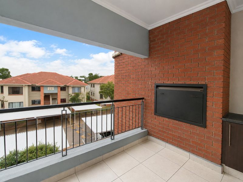 A covered balcony featuring a built-in braai, tiled flooring, and a metal balustrade, overlooking the apartment complex grounds.