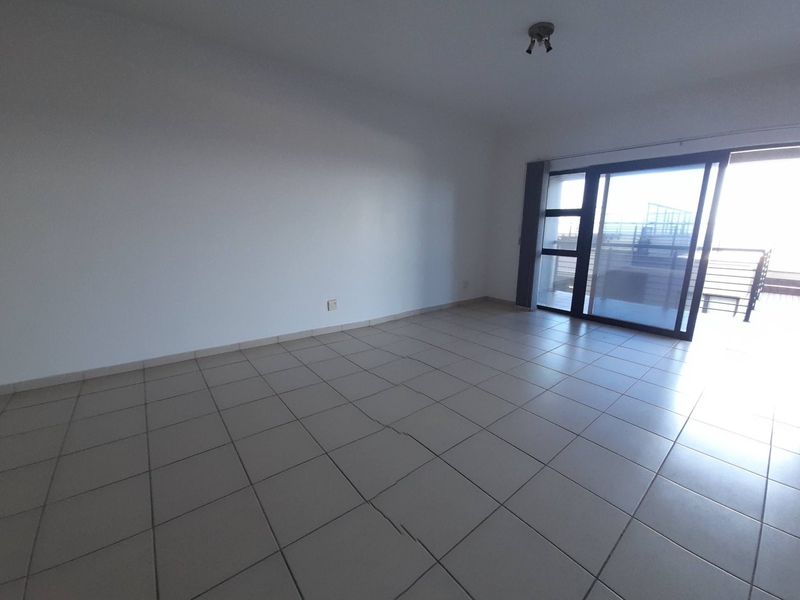 An empty, open-plan living area featuring neutral tiled flooring and a large sliding glass door providing access to an outdoor balcony.