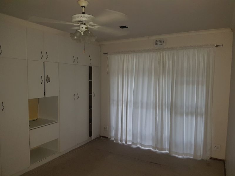 A vacant bedroom featuring extensive built-in cabinetry, a ceiling fan, and large window with curtains, requiring cosmetic updates to modernize the space.