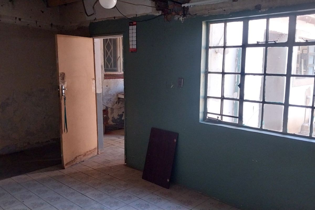 An empty living area featuring tiled flooring and steel-framed windows, currently requiring significant cosmetic renovation and wall repairs.