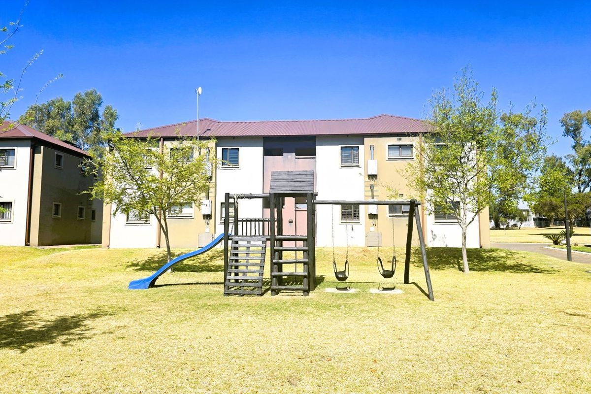 Exterior view of an apartment complex featuring a communal lawn area with a playground set and established trees.