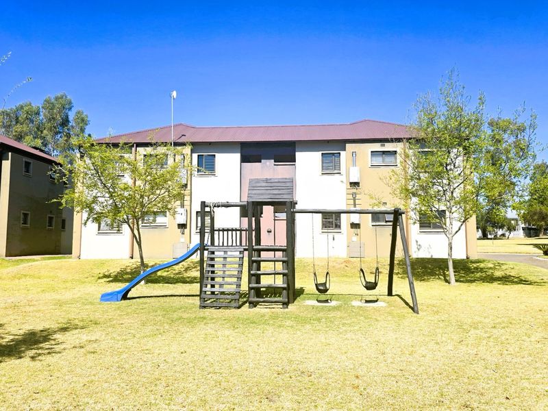 Exterior view of an apartment complex featuring a communal lawn area with a playground set and established trees.