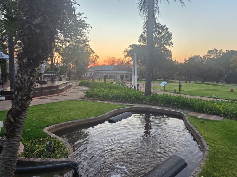A scenic view of a landscaped garden featuring a water feature, paved pathways, and timber decking overlooking a golf course at sunset.