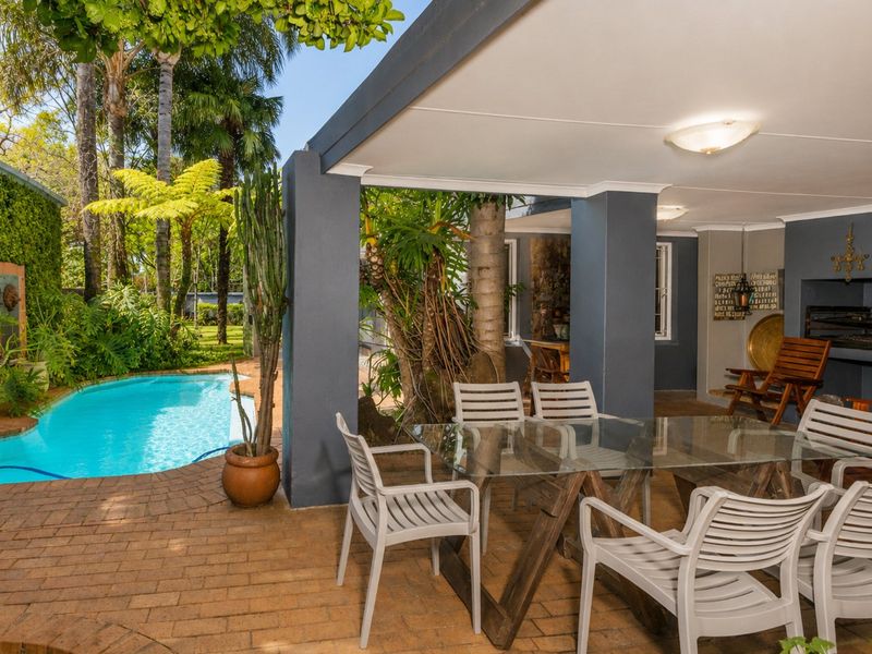 A covered outdoor patio area featuring a built-in braai, overlooking a swimming pool and lush garden setting.