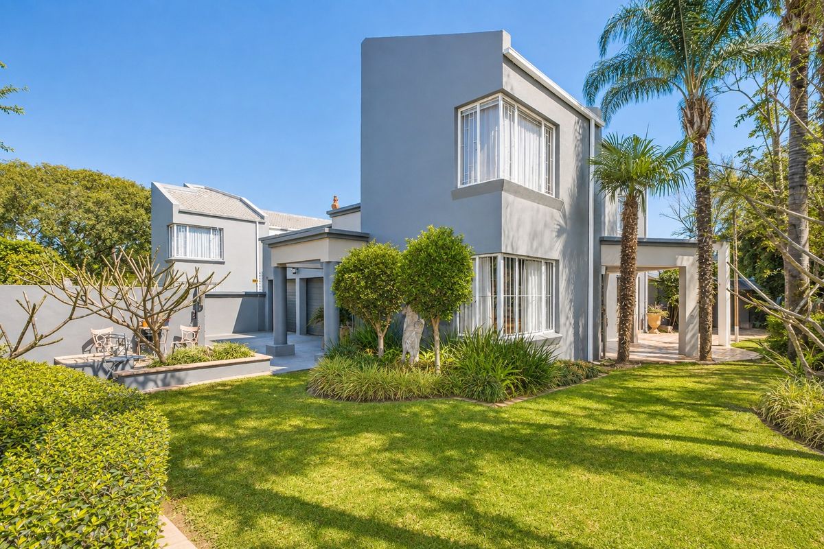 A well-maintained rear exterior view featuring a modern grey facade, paved patio area, and a lush landscaped garden with mature palm trees.