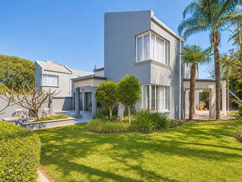 A well-maintained rear exterior view featuring a modern grey facade, paved patio area, and a lush landscaped garden with mature palm trees.