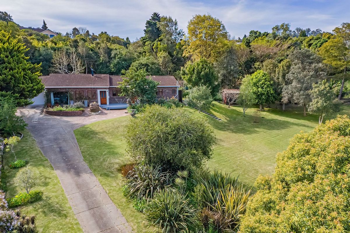 Aerial view of a brick-faced house featuring a spacious lawn, mature trees, and a long concrete driveway set within a lush, landscaped garden.