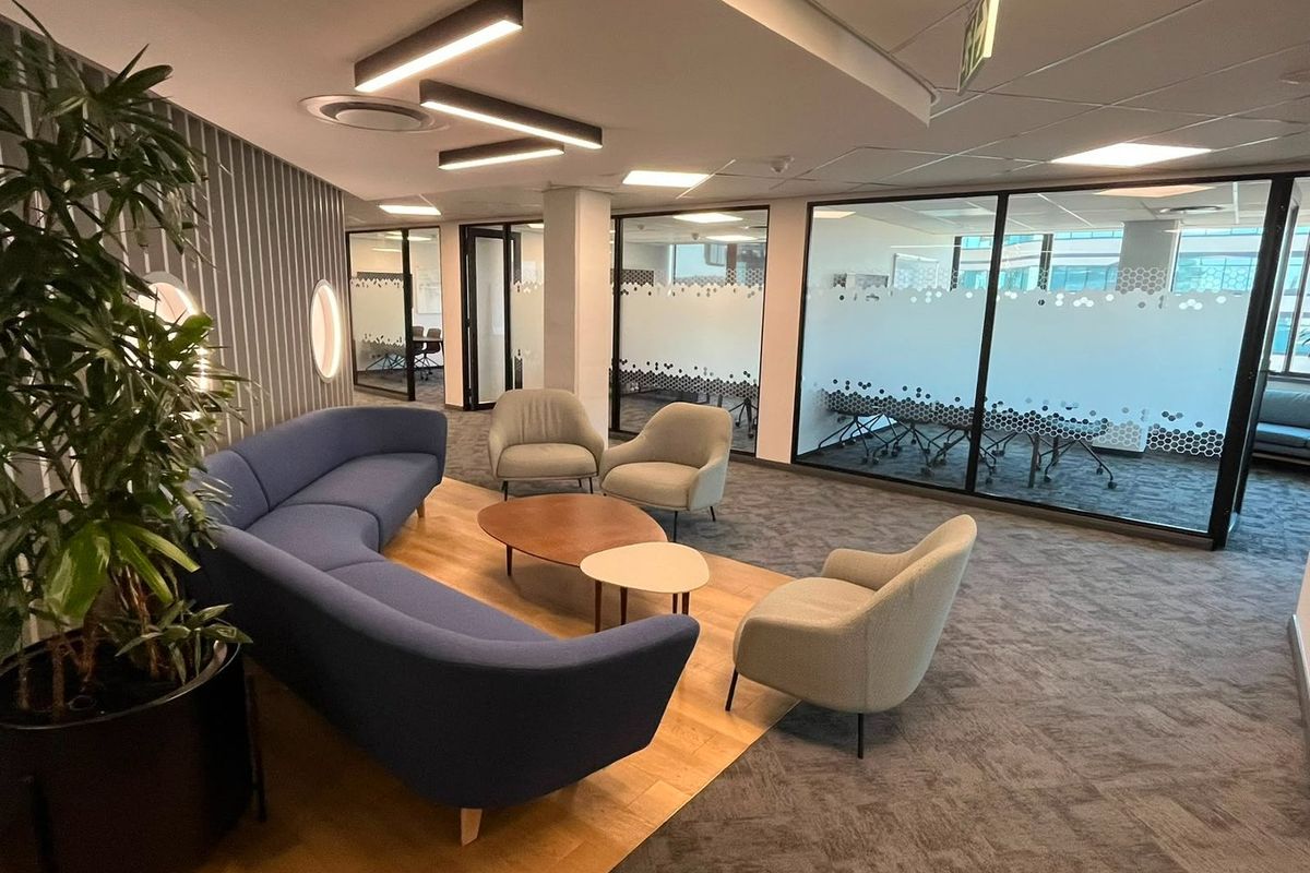 Modern commercial lounge area featuring glass-partitioned meeting rooms, contemporary linear lighting, and a mix of carpet and wood flooring in a professional office setting.
