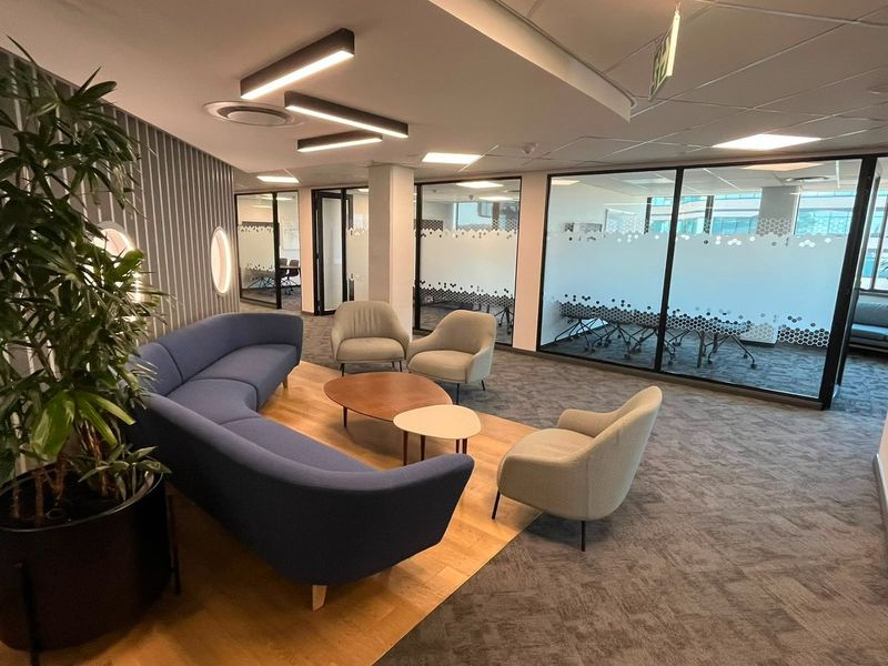 Modern commercial lounge area featuring glass-partitioned meeting rooms, contemporary linear lighting, and a mix of carpet and wood flooring in a professional office setting.