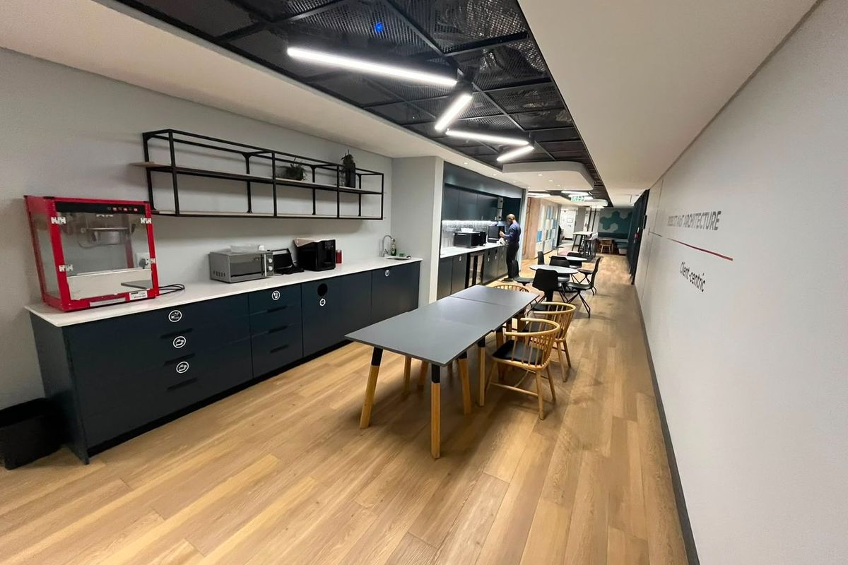 Modern commercial kitchen and break area featuring sleek dark cabinetry, integrated countertops, exposed mesh ceiling, and contemporary linear lighting.