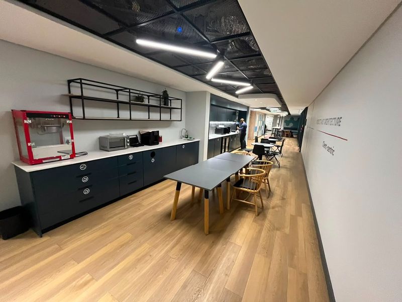 Modern commercial kitchen and break area featuring sleek dark cabinetry, integrated countertops, exposed mesh ceiling, and contemporary linear lighting.