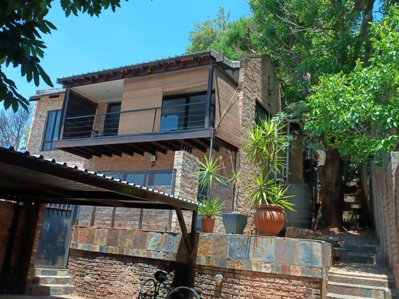 Rear view of a multi-level house featuring exposed brickwork, a timber balcony with metal railings, and stone retaining walls set against a natural backdrop.