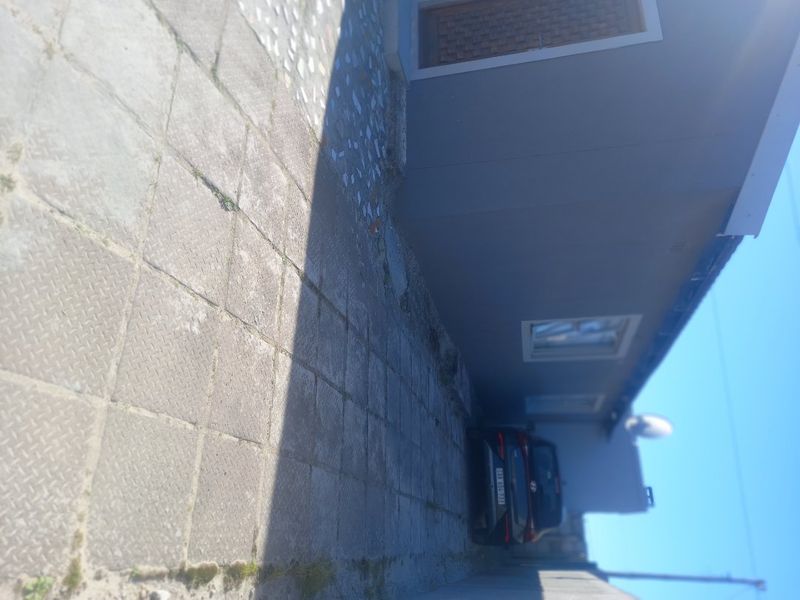 A side or rear exterior view of the property featuring paved ground and a grey rendered wall with a window and door.